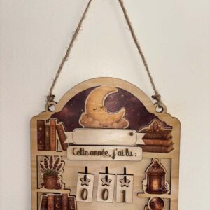 Compteur de lecture en bois personnalisable représentant une lune endormie entourée de livres, d'une lanterne et d'éléments inspirés des univers fantastiques. Les chiffres amovibles permettent de suivre facilement les lectures réalisées au cours de l'année.