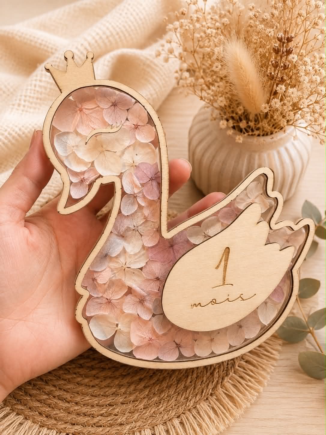 Carte étape bébé en bois en forme de cygne personnalisée avec prénom, décorée de fleurs séchées et équipée d'ailes interchangeables indiquant les mois de bébé.