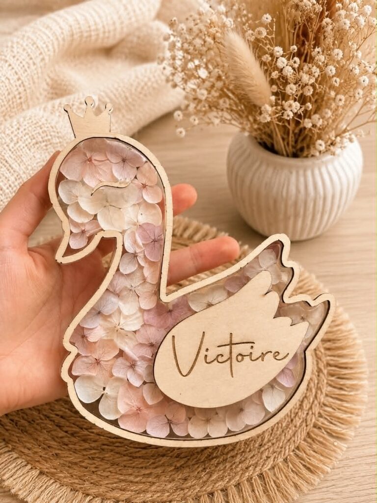 Carte étape bébé en bois en forme de cygne personnalisée avec prénom, décorée de fleurs séchées et équipée d'ailes interchangeables indiquant les mois de bébé.