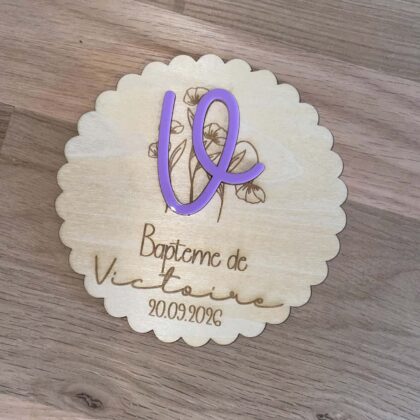 Plaque de baptême en bois naturel personnalisée avec le prénom de l'enfant et la date du baptême, avec un prénom en acrylique violet en relief et un motif floral gravé.