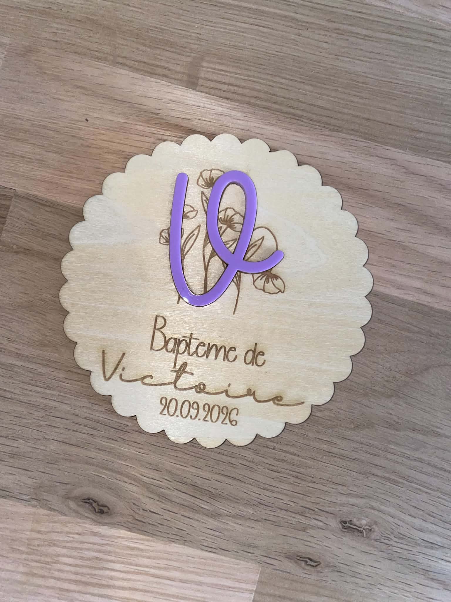 2d9c0dd6 a532 4454 a01d 76b2dcc23a8e Plaque de baptême en bois naturel personnalisée avec le prénom de l'enfant et la date du baptême, avec un prénom en acrylique violet en relief et un motif floral gravé.